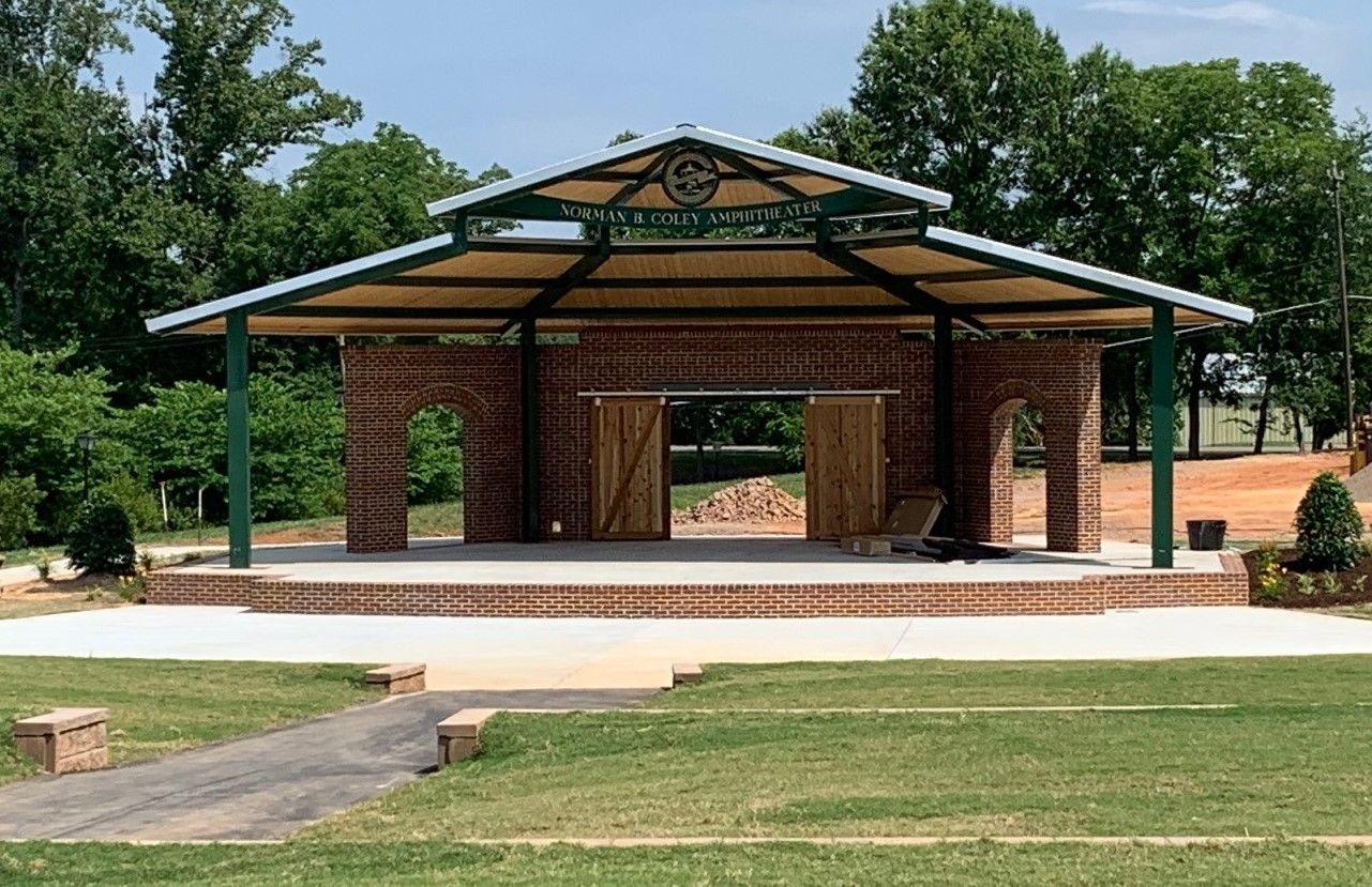 Open-air brick pavilion in a park with a green metal roof and trees in the background
