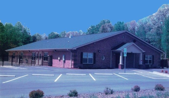 Single-story brick building with a covered entrance and empty parking lot, surrounded by trees