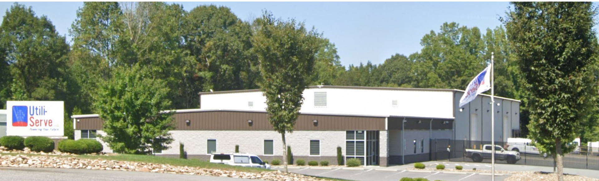 Low brick building with a white roof, flagpoles, and a roadside sign, surrounded by trees and parked cars