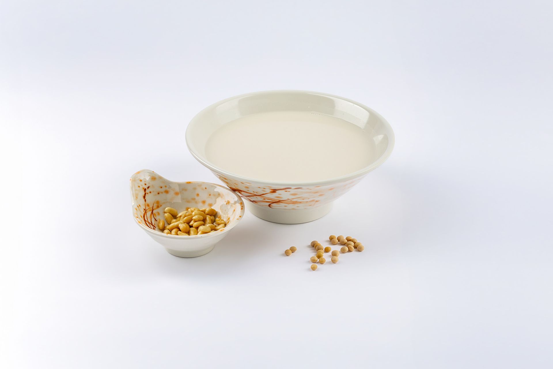 A bowl of light-colored soy milk served with pine nuts in a small decorative bowl and scattered on a white background.