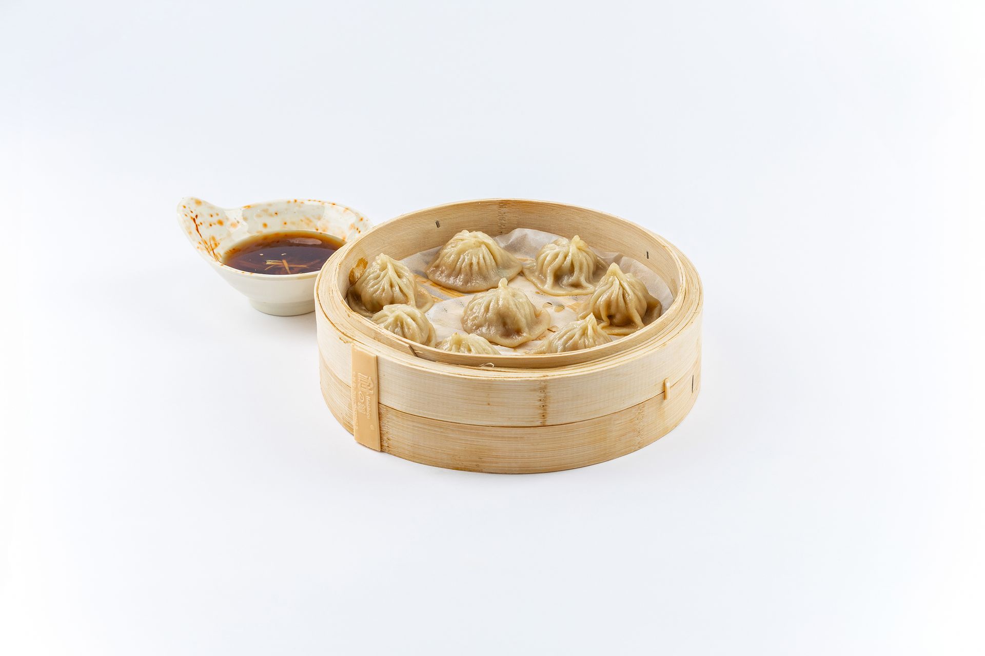 A bamboo steamer basket filled with six steamed soup dumplings next to a small dish of soy dipping sauce.