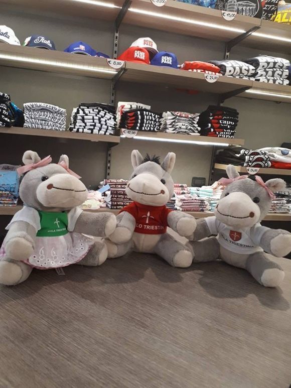 Tre peluche a forma di asino con fiocchi, ognuno con una maglietta decorata, su un tavolo in un negozio.