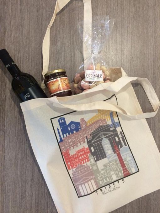 Borsa tote con disegno dello skyline di Trieste, contenente una bottiglia di vino, un barattolo di marmellata e biscotti confezionati.