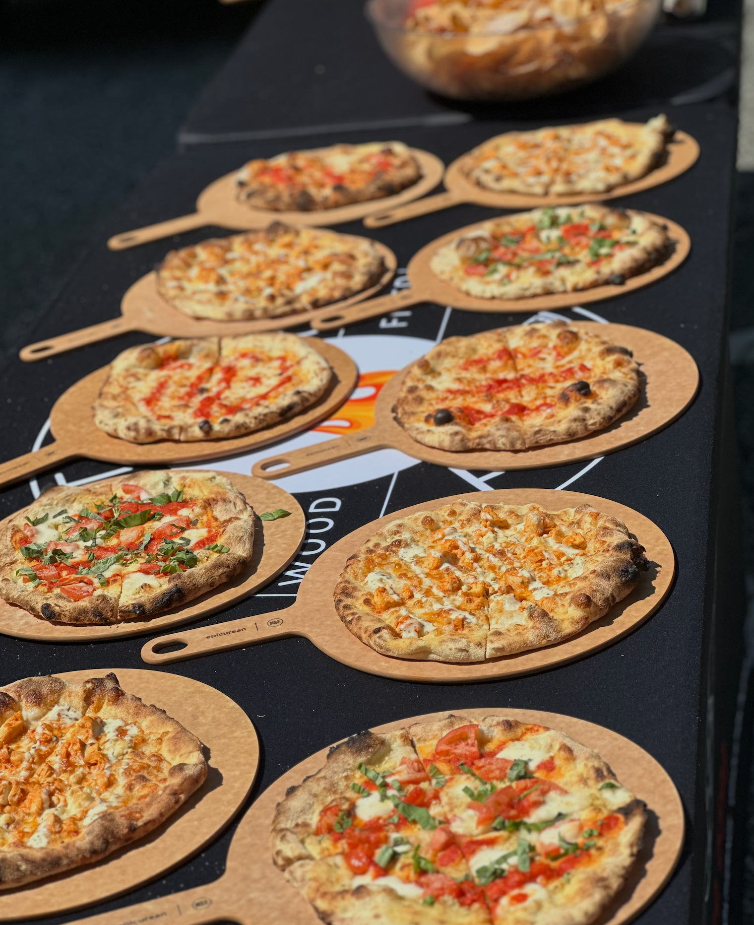 Several pizzas on wooden paddles displayed on a black table at an outdoor setting.