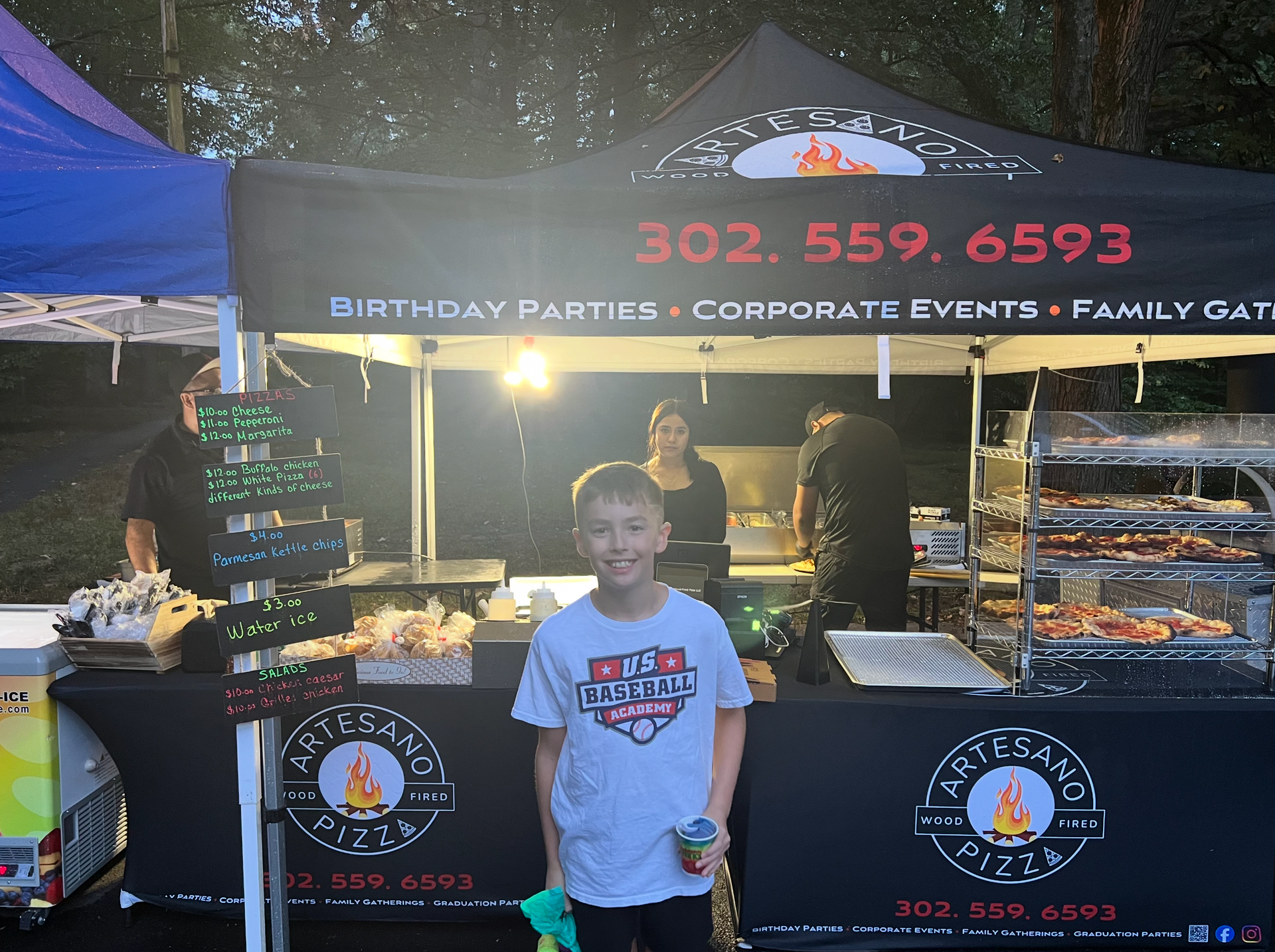 Boy in front of a food stand with people, sign reads 