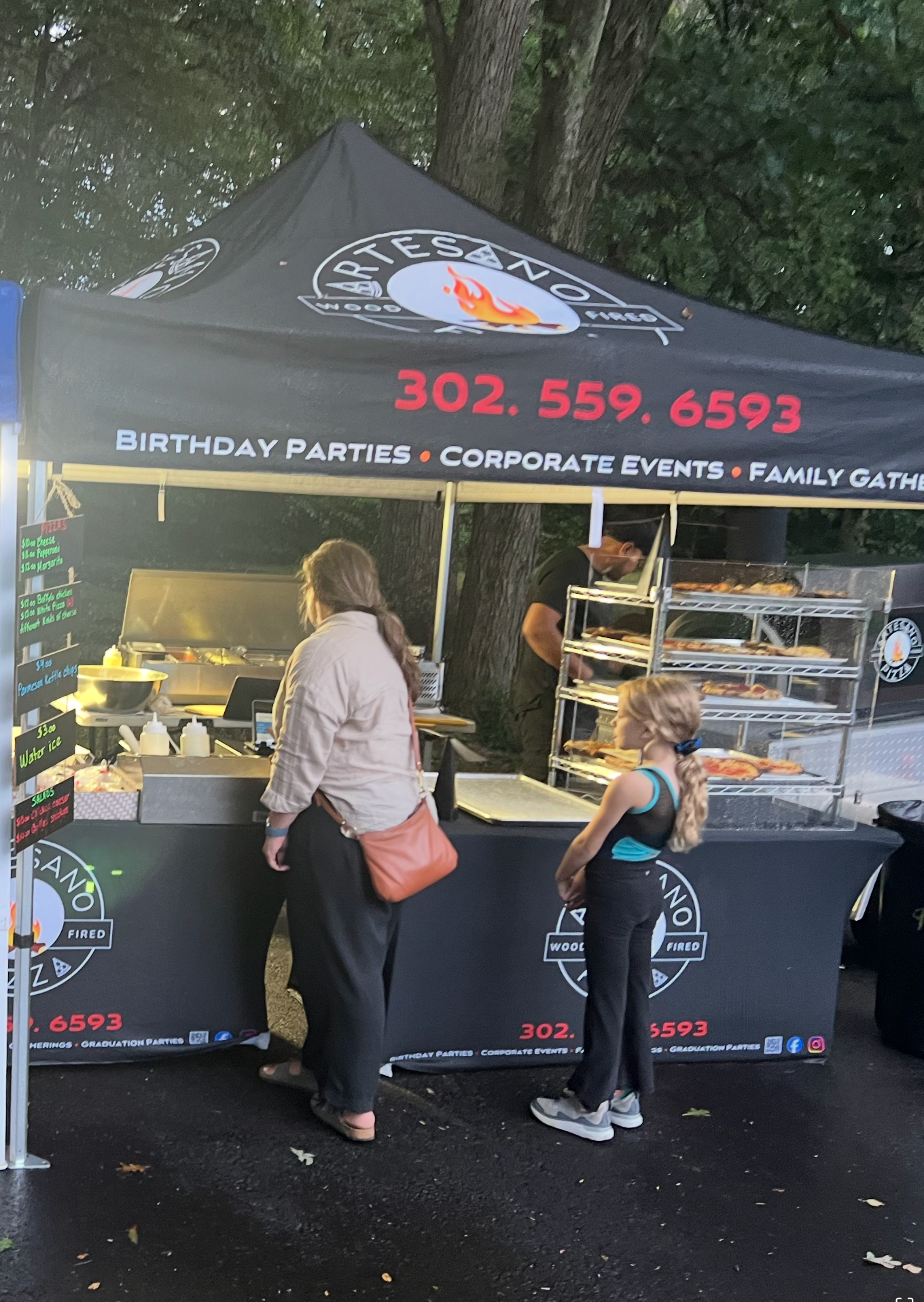 A food vendor tent with a woman and a child looking at the display, the vendor is 302-559-6593.