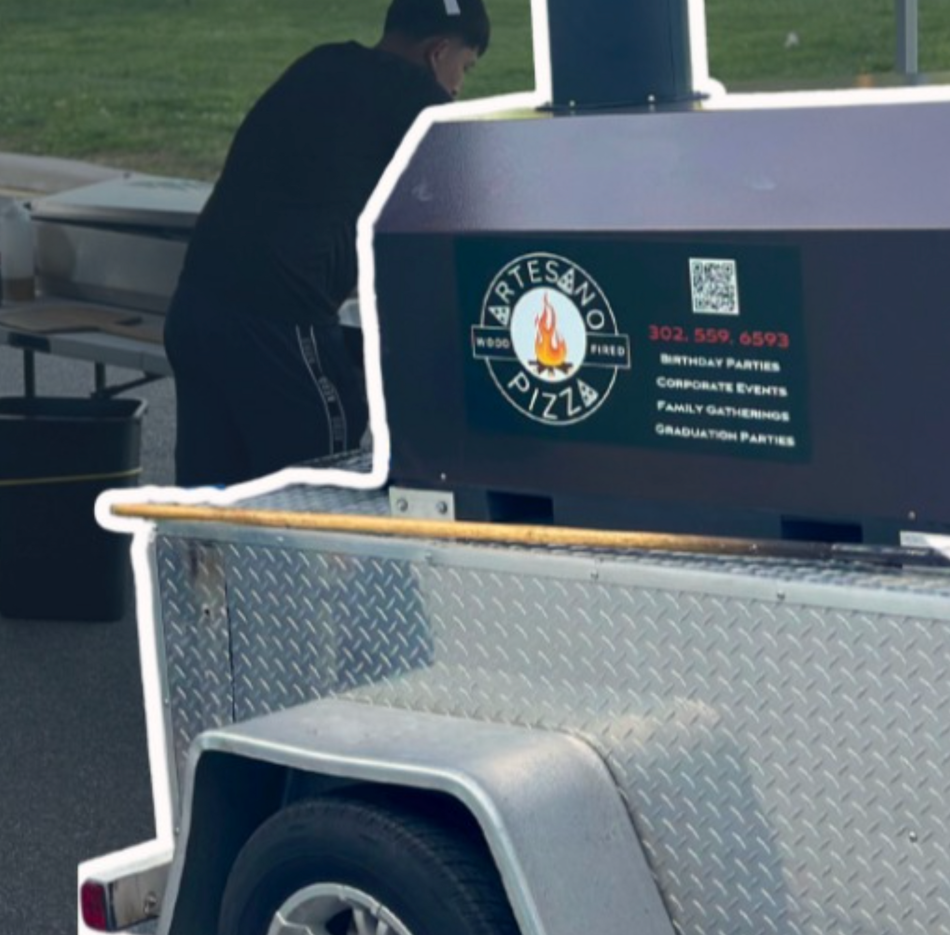 Man at Artesano Pizza food trailer. Trailer is silver, with a dark purple pizza oven.