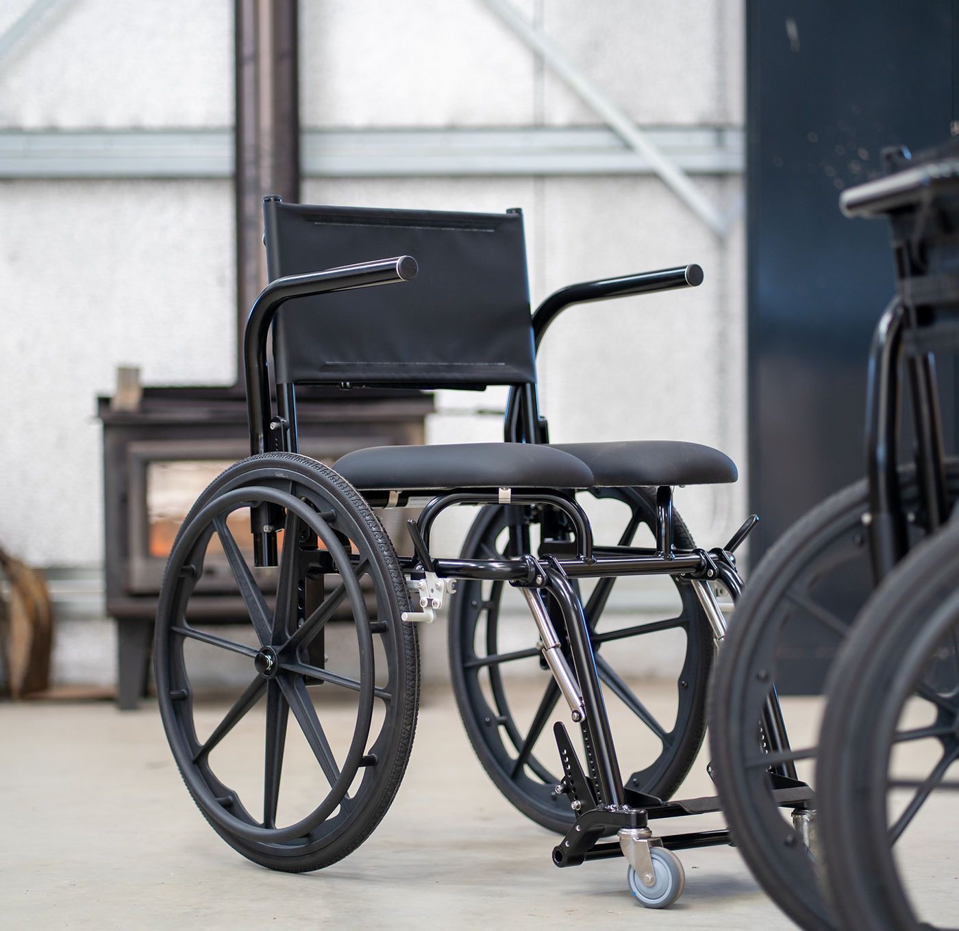 Black wheelchair with armrests, in a workshop setting with a wood-burning stove visible.