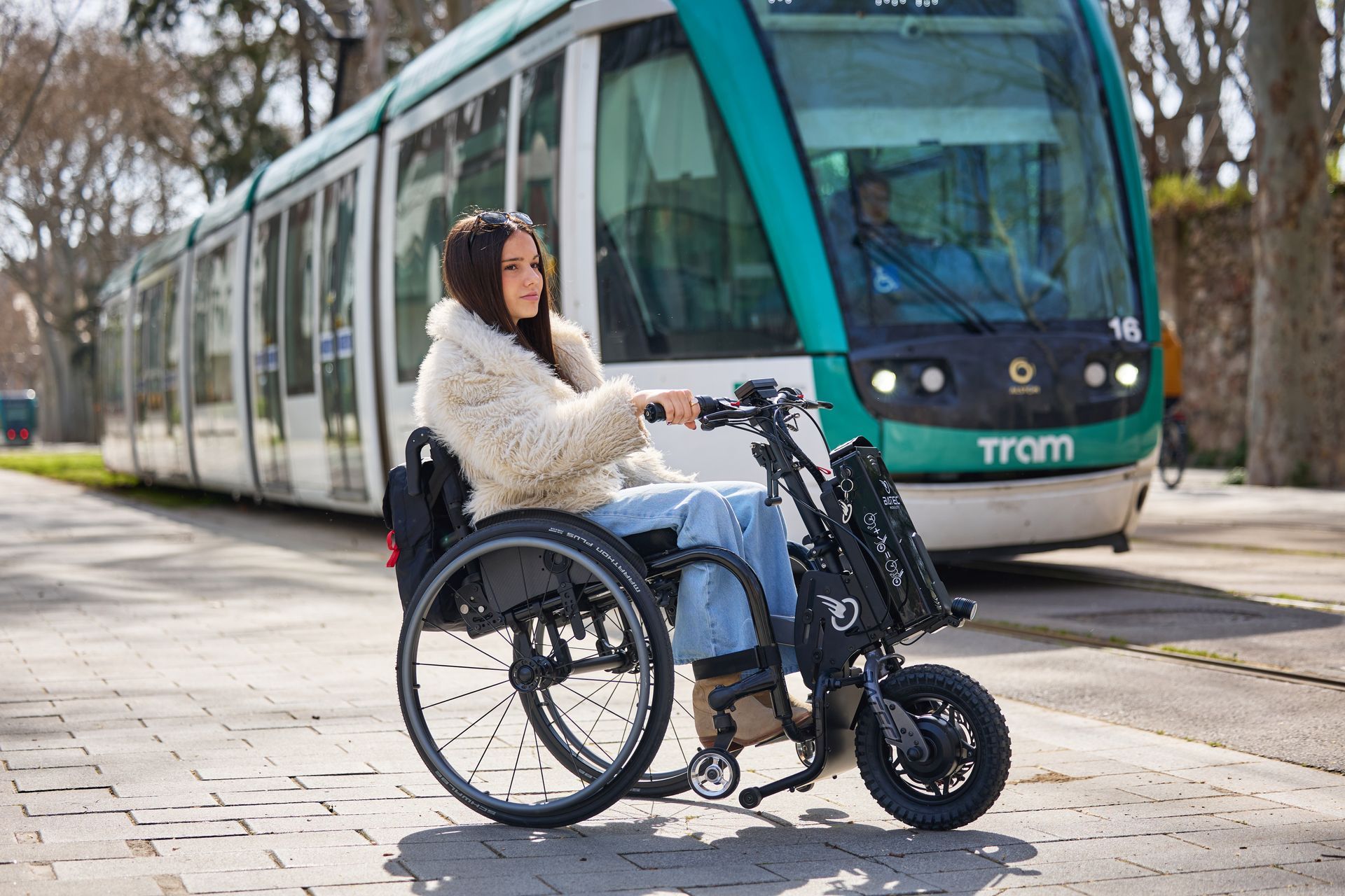 Woman in a wheelchair on a sidewalk beside a green tram in a city street