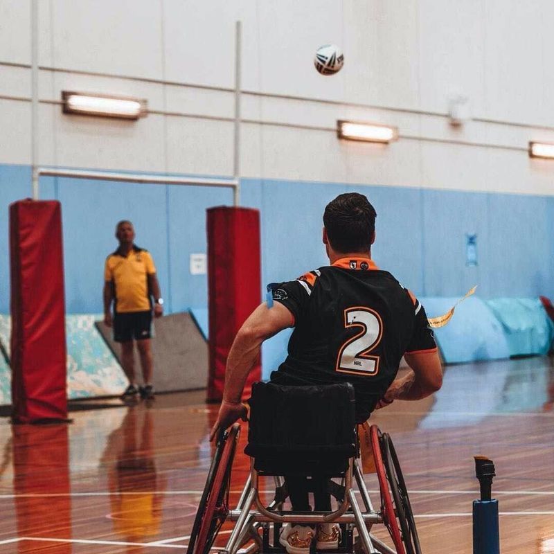 Man in wheelchair throwing a ball indoors; another man watches.