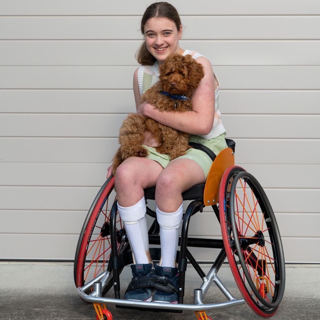 Smiling girl in wheelchair holding brown puppy; outdoors by a wall.