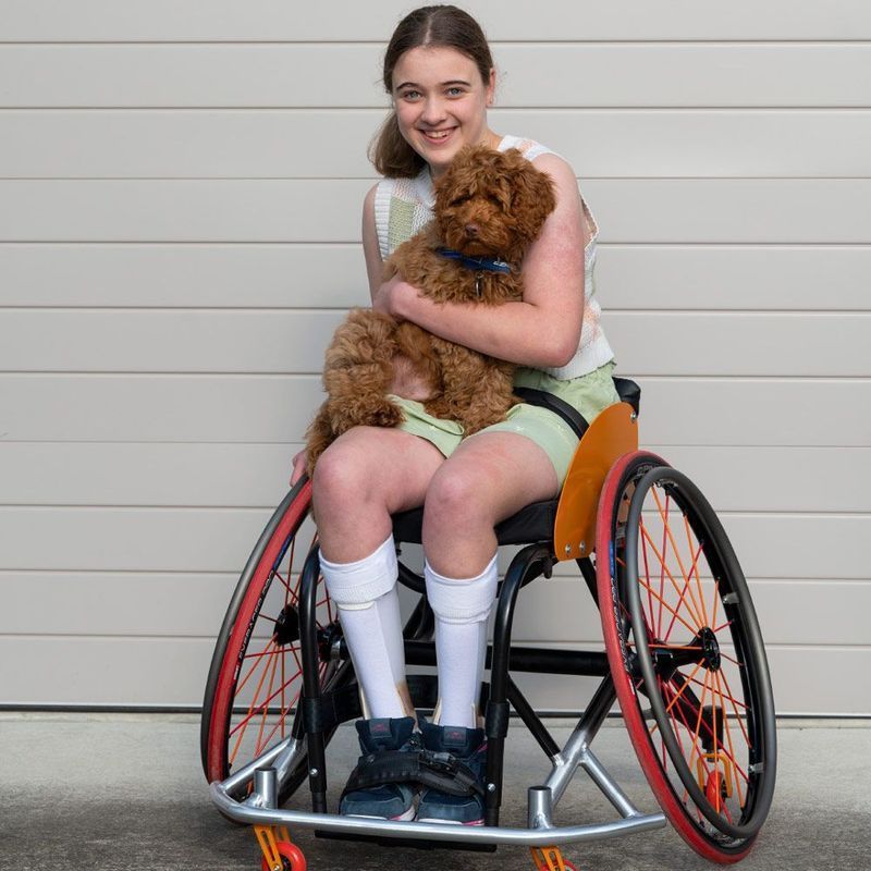 Teenage girl in a wheelchair smiles, holding a small brown dog in front of a garage door.