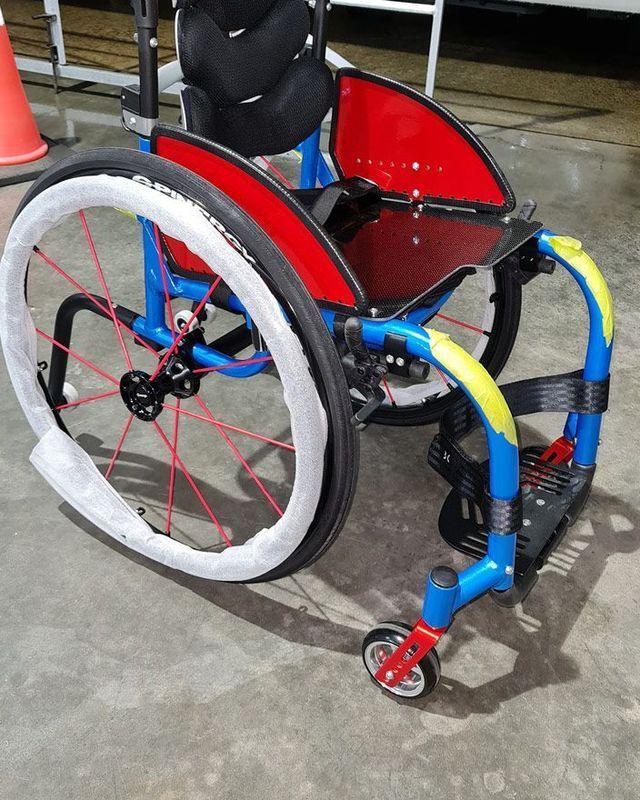 Blue, red, and yellow racing wheelchair with protective white wheel covers, set on a gray surface.