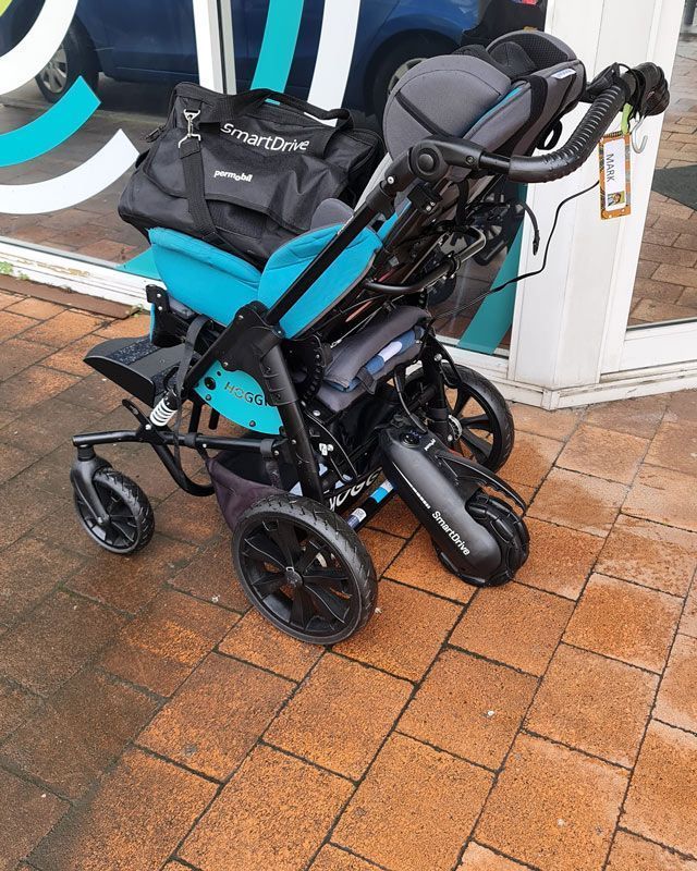Special needs stroller with turquoise seat, black frame, and wheels, on brick sidewalk.