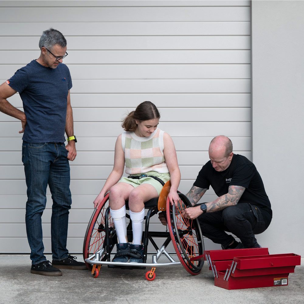 Two men and a young woman in a wheelchair. Man fixes wheel. White wall backdrop.