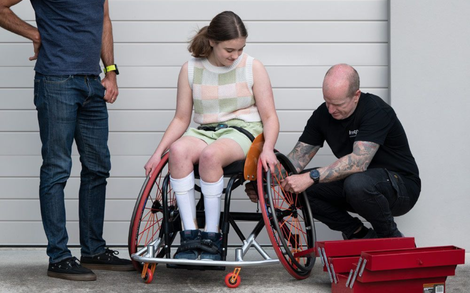 Woman in wheelchair with two men; one adjusts her wheel. Outdoors, garage.
