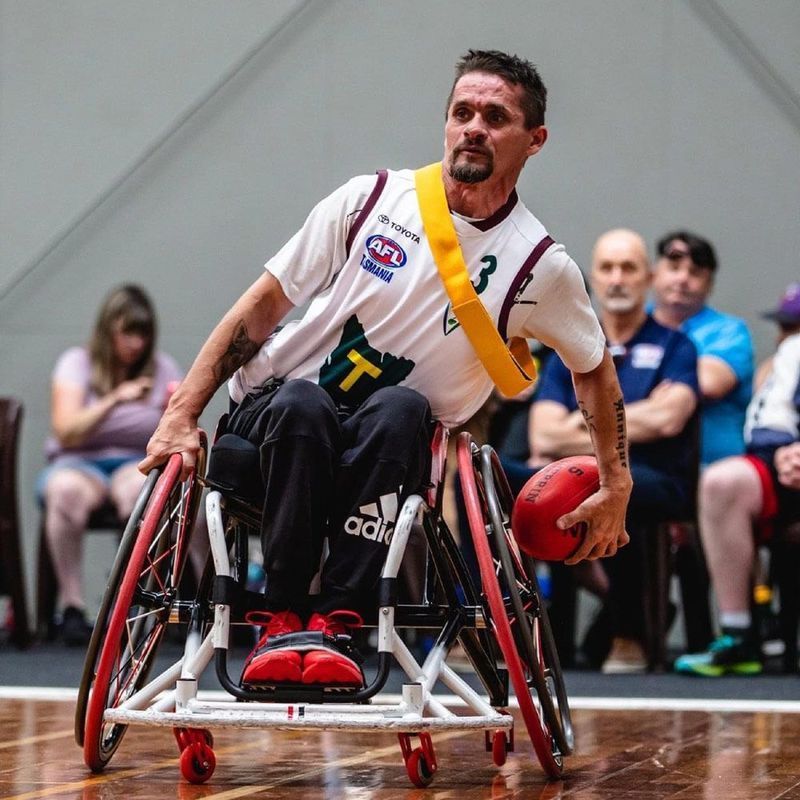 Man in wheelchair playing wheelchair football, indoor court, holding ball, spectators in background.