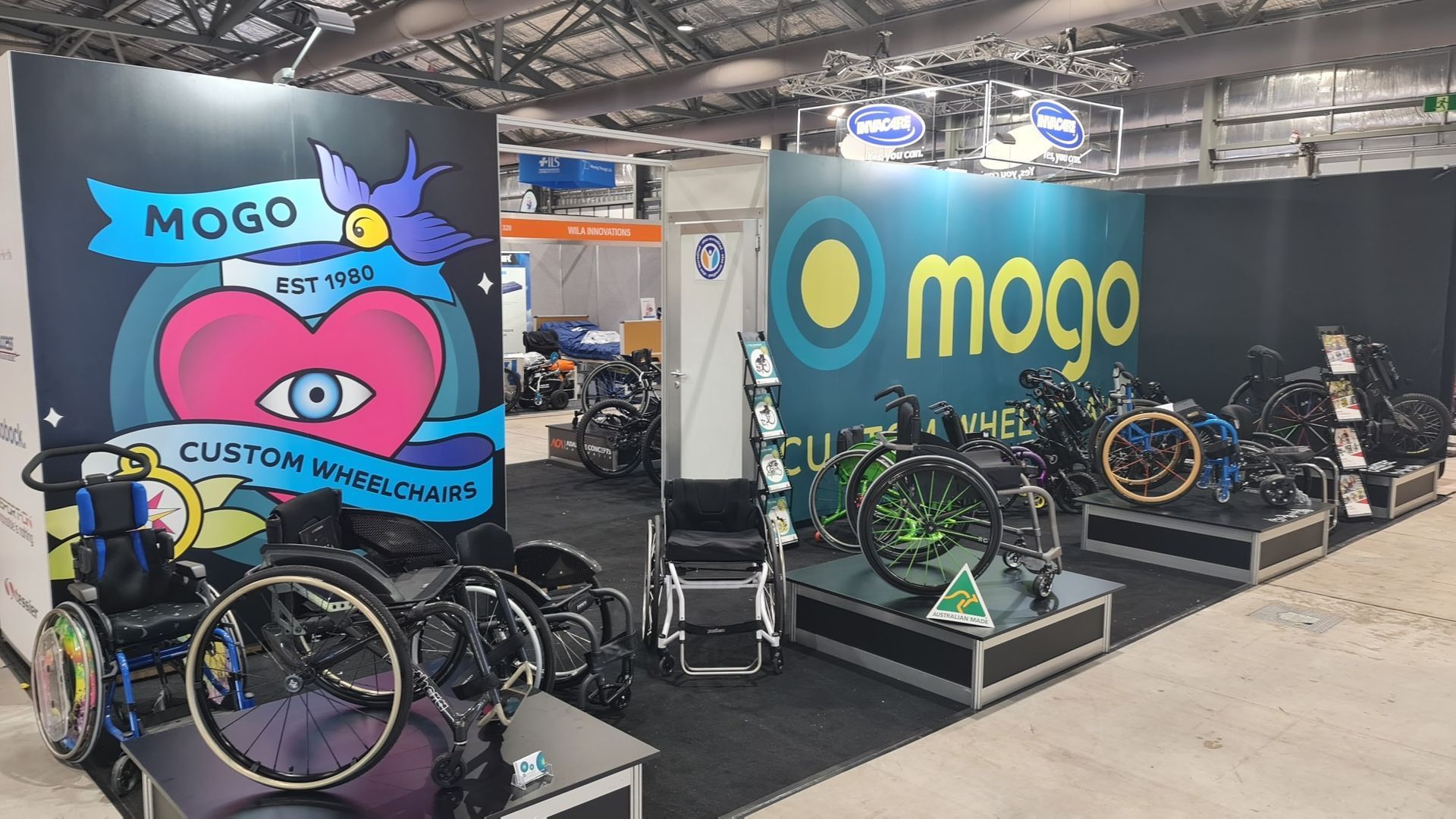 Mogo wheelchair display at a trade show. Wheelchairs in various colors are displayed with signage and branding.