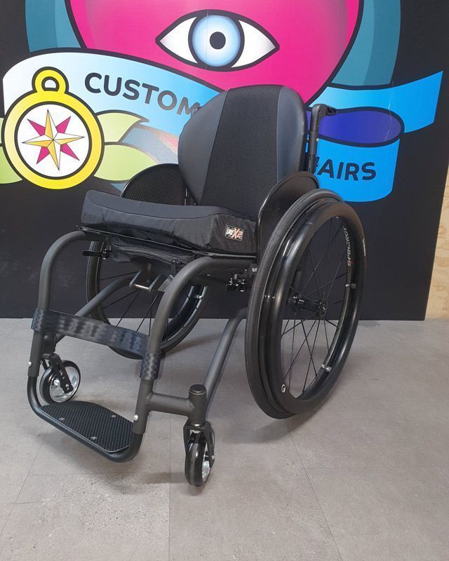 Black wheelchair with gray seat and back, on a gray floor, in front of a colorful sign.