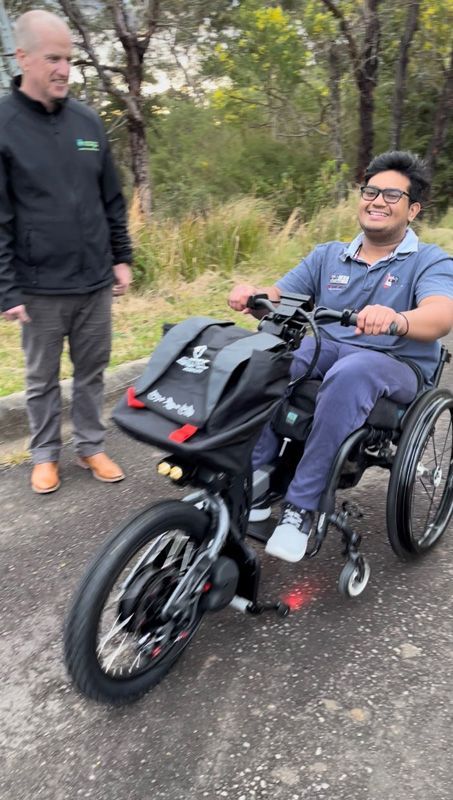Man in wheelchair with electric assist smiling, riding on pavement, another man watches.