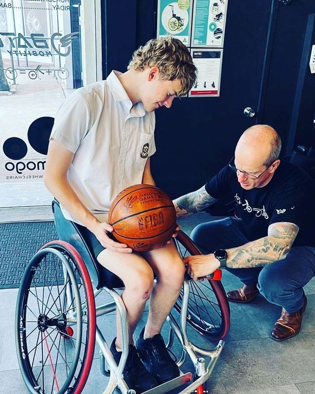 Man in wheelchair holding basketball; another man adjusts the wheelchair's wheel. Indoors, in front of a shop.