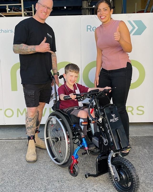 Man and woman giving thumbs up with a smiling boy in a wheelchair in front of a business sign.