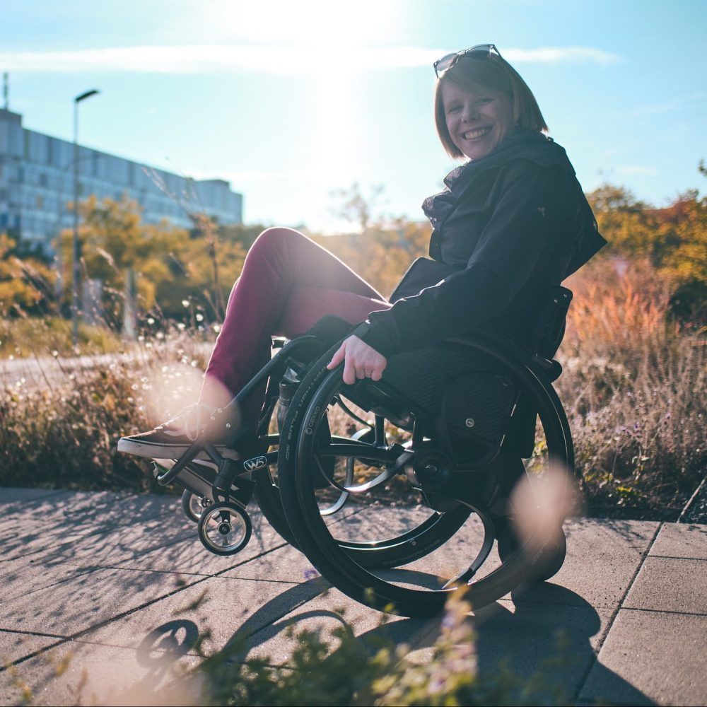 Woman in wheelchair smiles outdoors, sunny day.