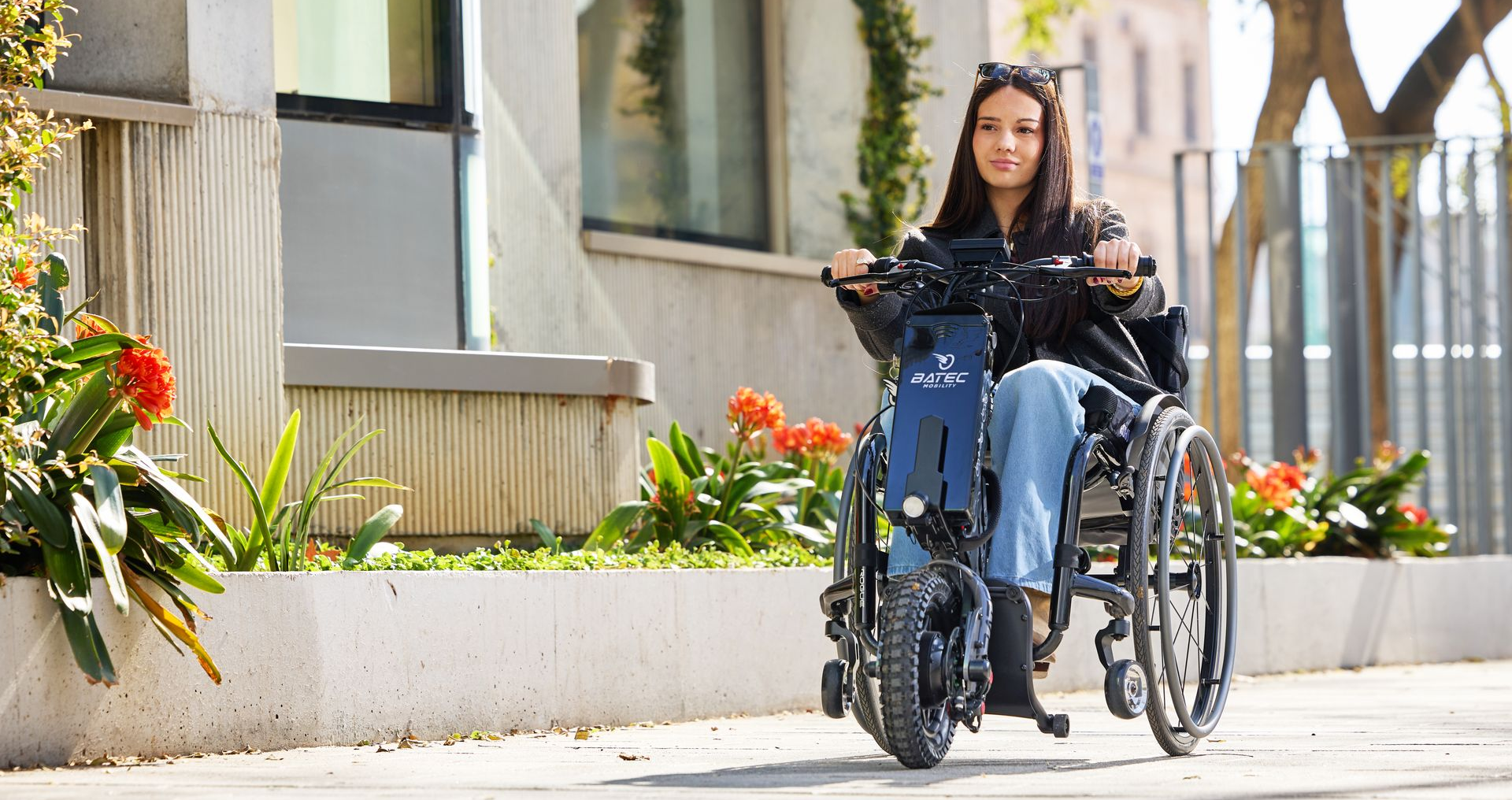 Woman in a wheelchair riding along a sunlit sidewalk beside a building and flower bed