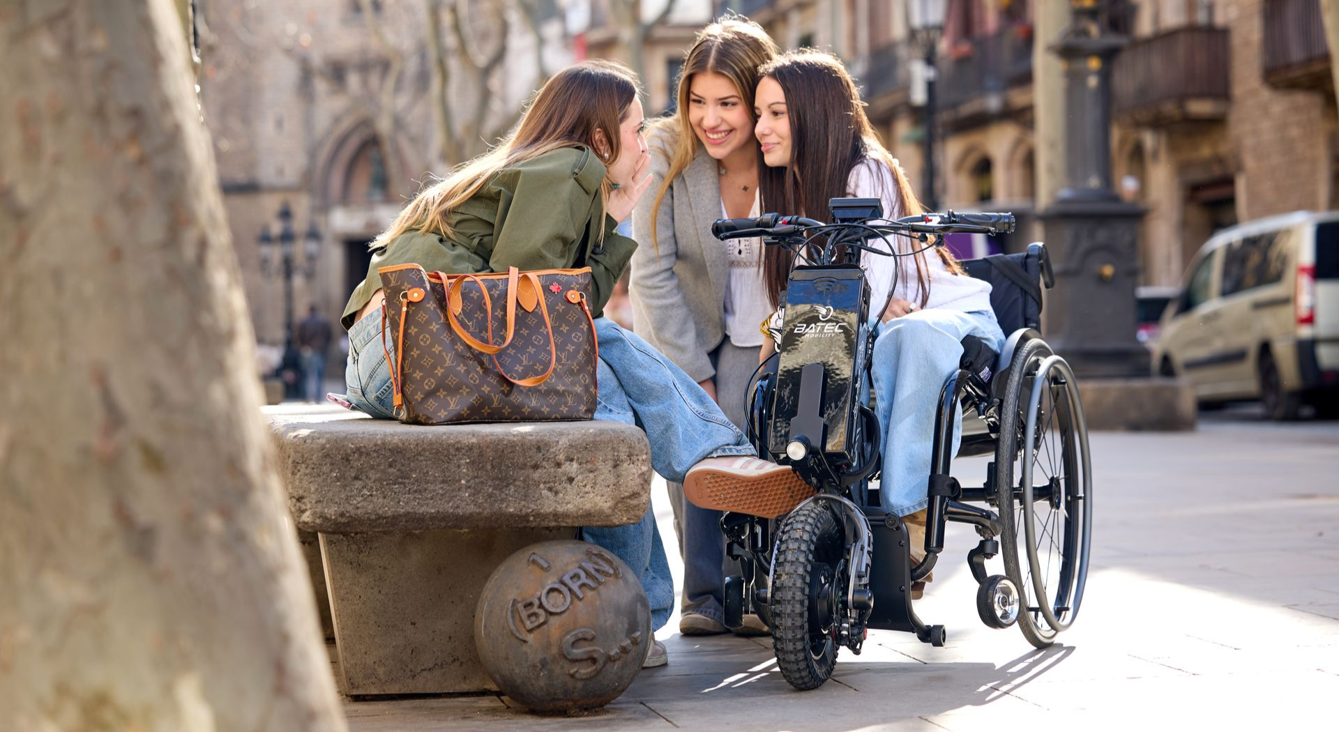 Two people sit beside a wheelchair on a sunlit cobblestone street, smiling near a parked bicycle.