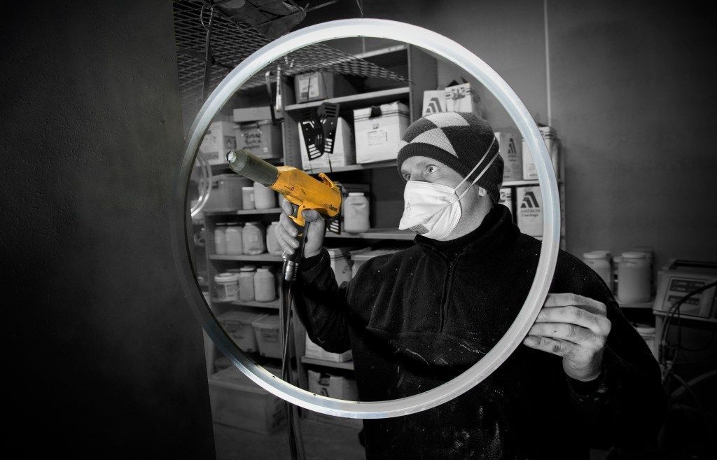 Person spray painting a metal rim with a yellow tool, wearing a respirator, in a workshop.
