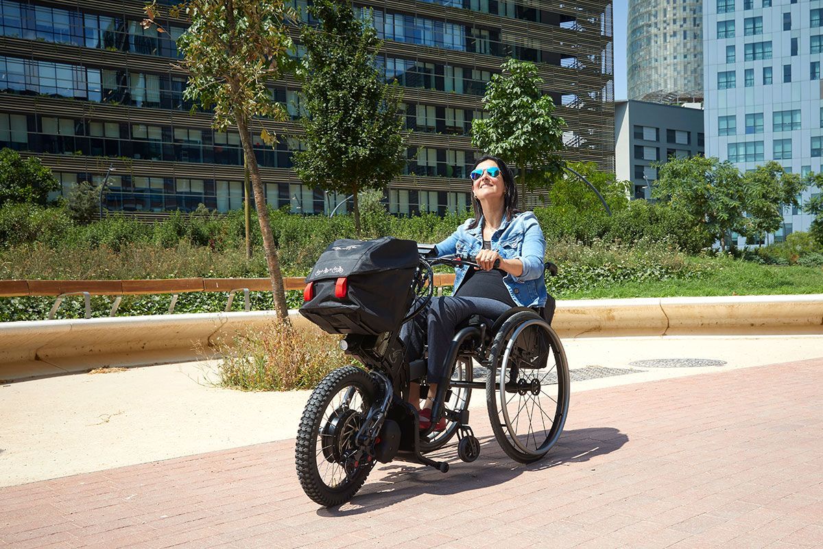 Person seated on a wheelchair-bike in a sunny urban park walkway with modern buildings behind.