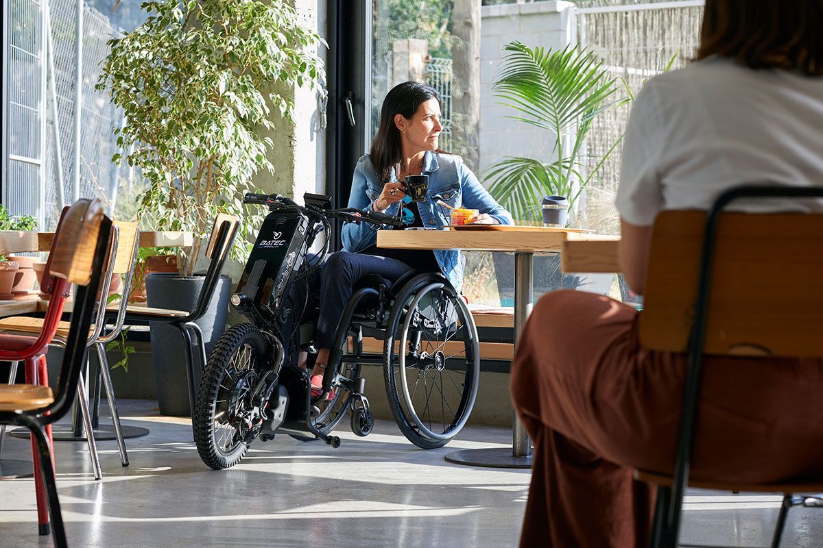 Person in a wheelchair sitting in a bright café, speaking with another seated person near a window.