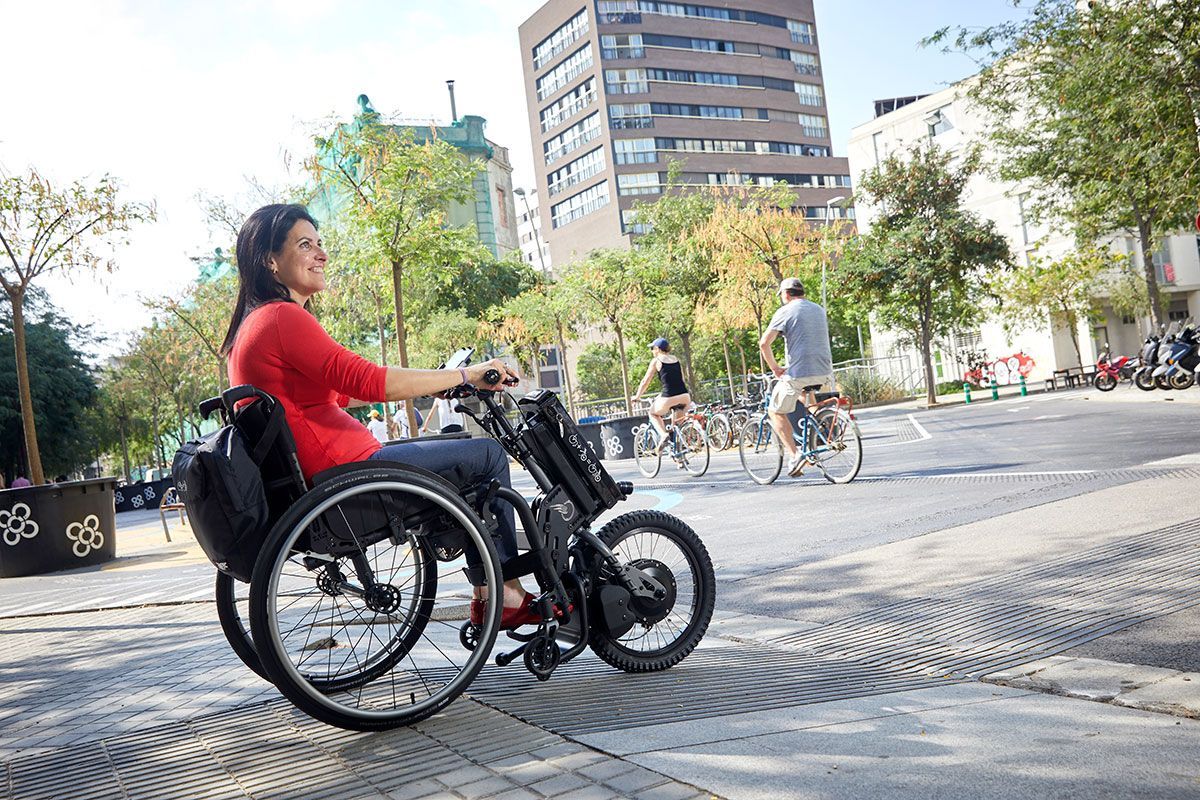 Person in a wheelchair with a front bike attachment riding in a city plaza with cyclists and buildings.