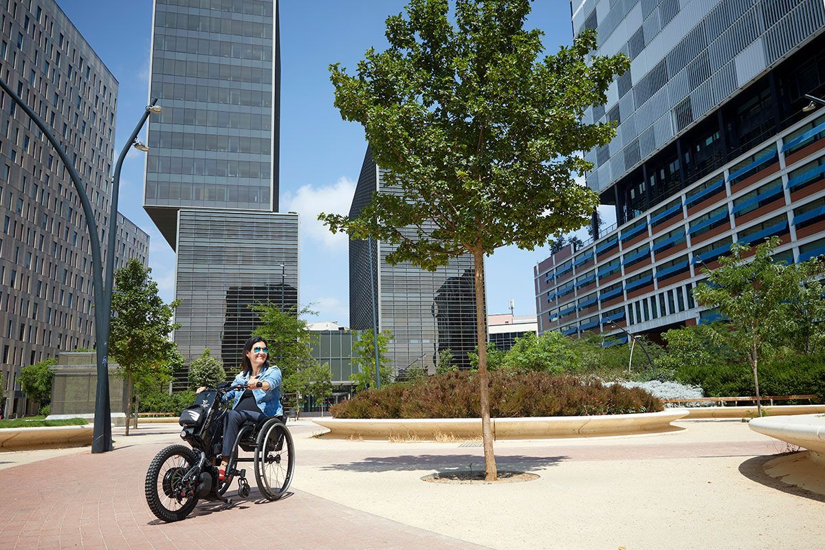 Two people in a wheelchair in a modern city plaza with trees and tall glass buildings.