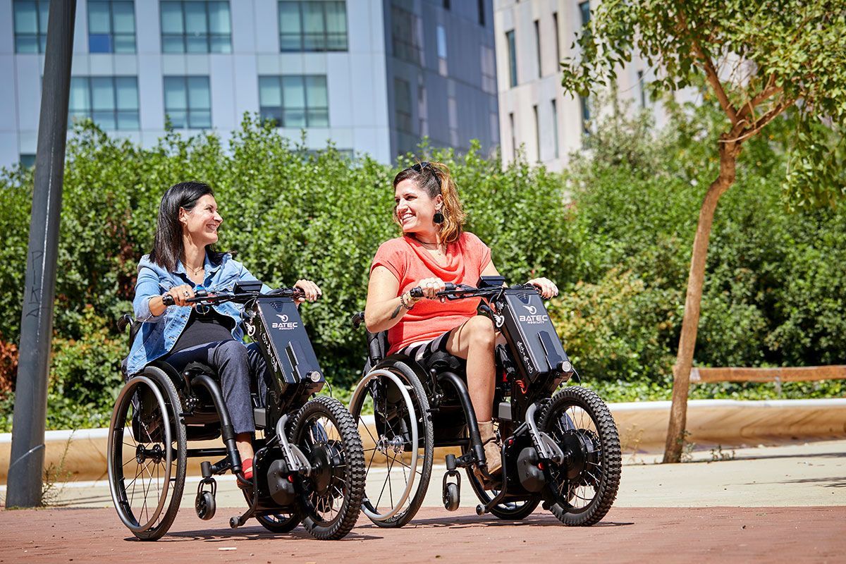 Two people in wheelchairs talking outdoors on a city sidewalk with trees and buildings behind them