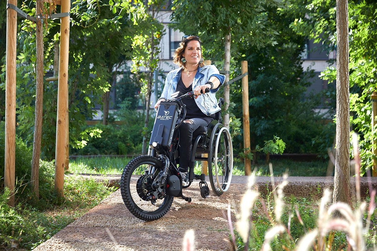 Person in a wheelchair outdoors on a sunny path with green trees and grass.