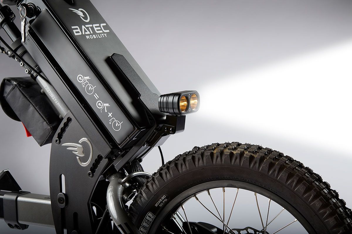 Close-up of a black Batec mobility aid with a lit headlight and a rear wheel with a knobby tire.