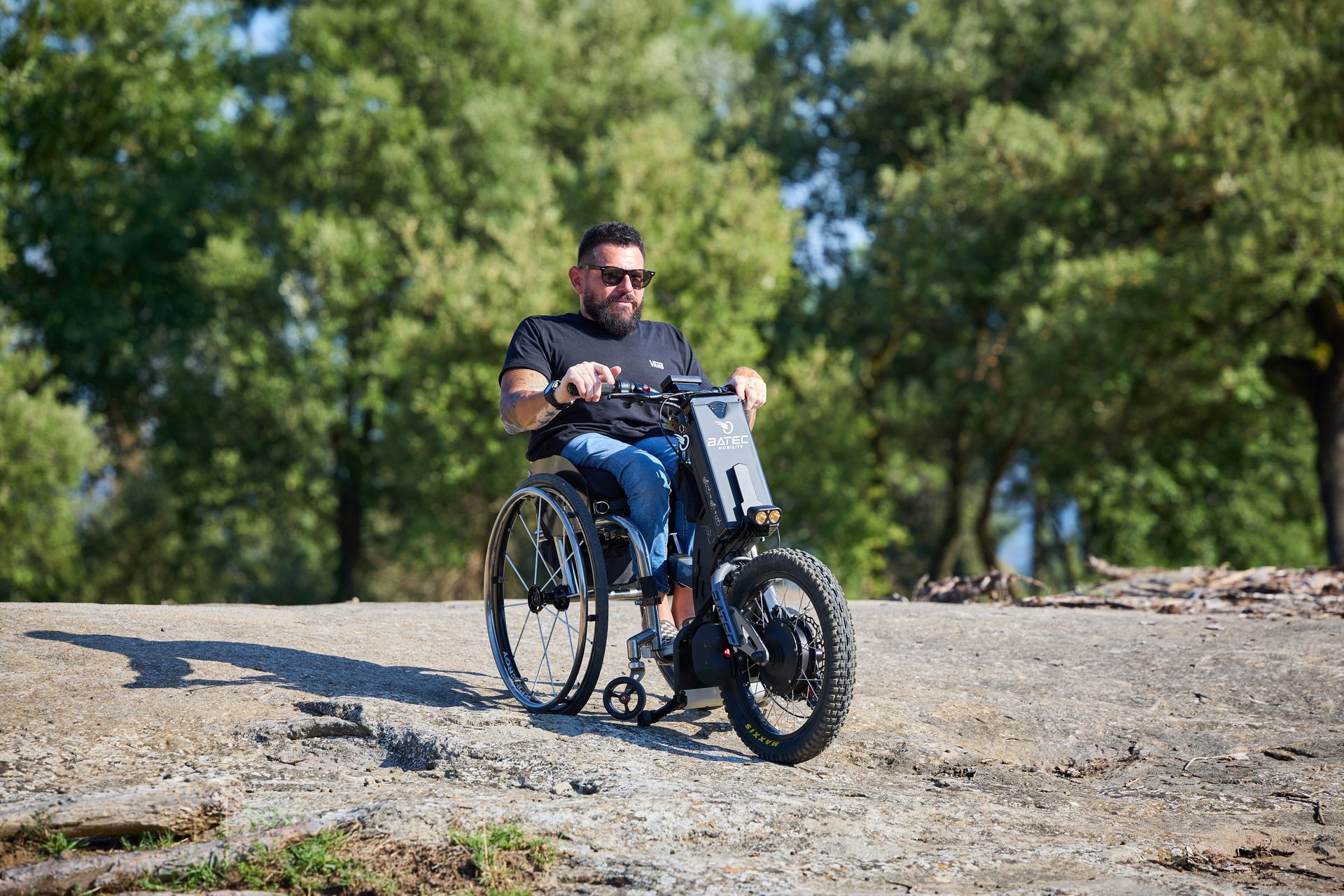 Man in a wheelchair rides on a gravel path in a sunny park, with trees in the background.