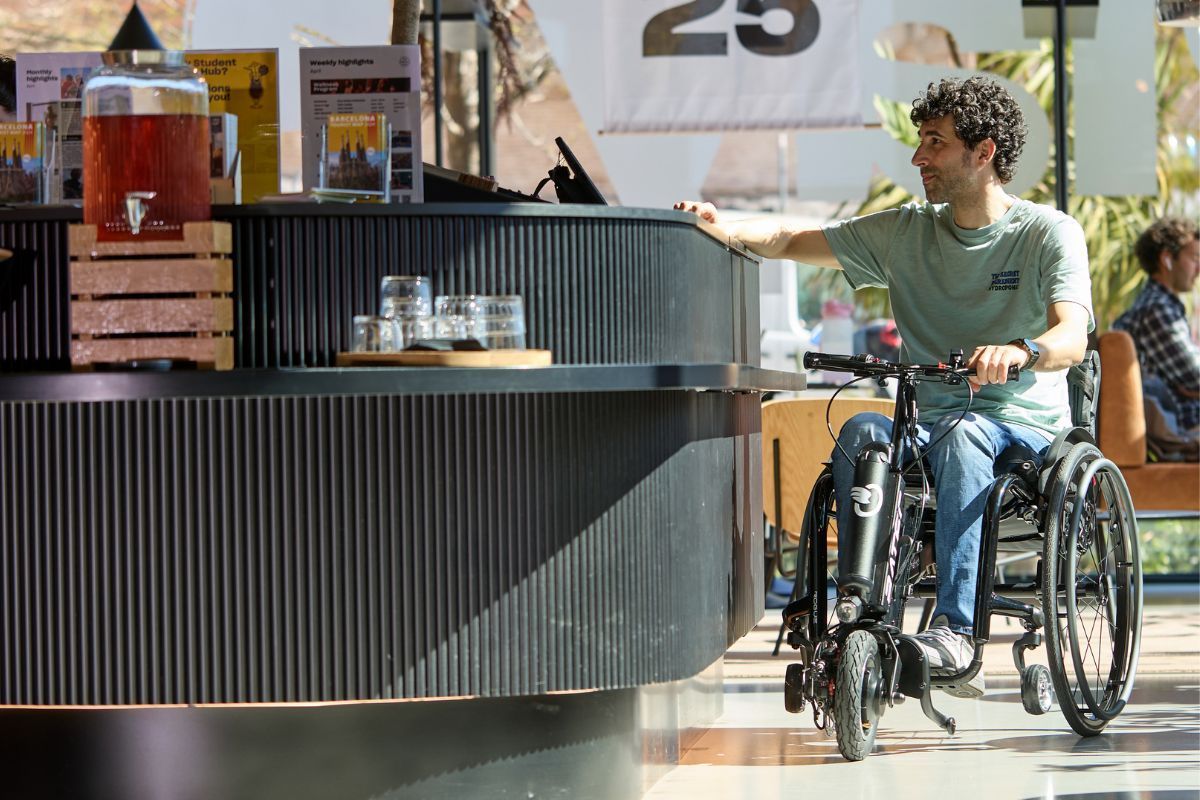 Man in wheelchair at a counter. Reaches towards the counter with beverage dispenser and glasses on it.