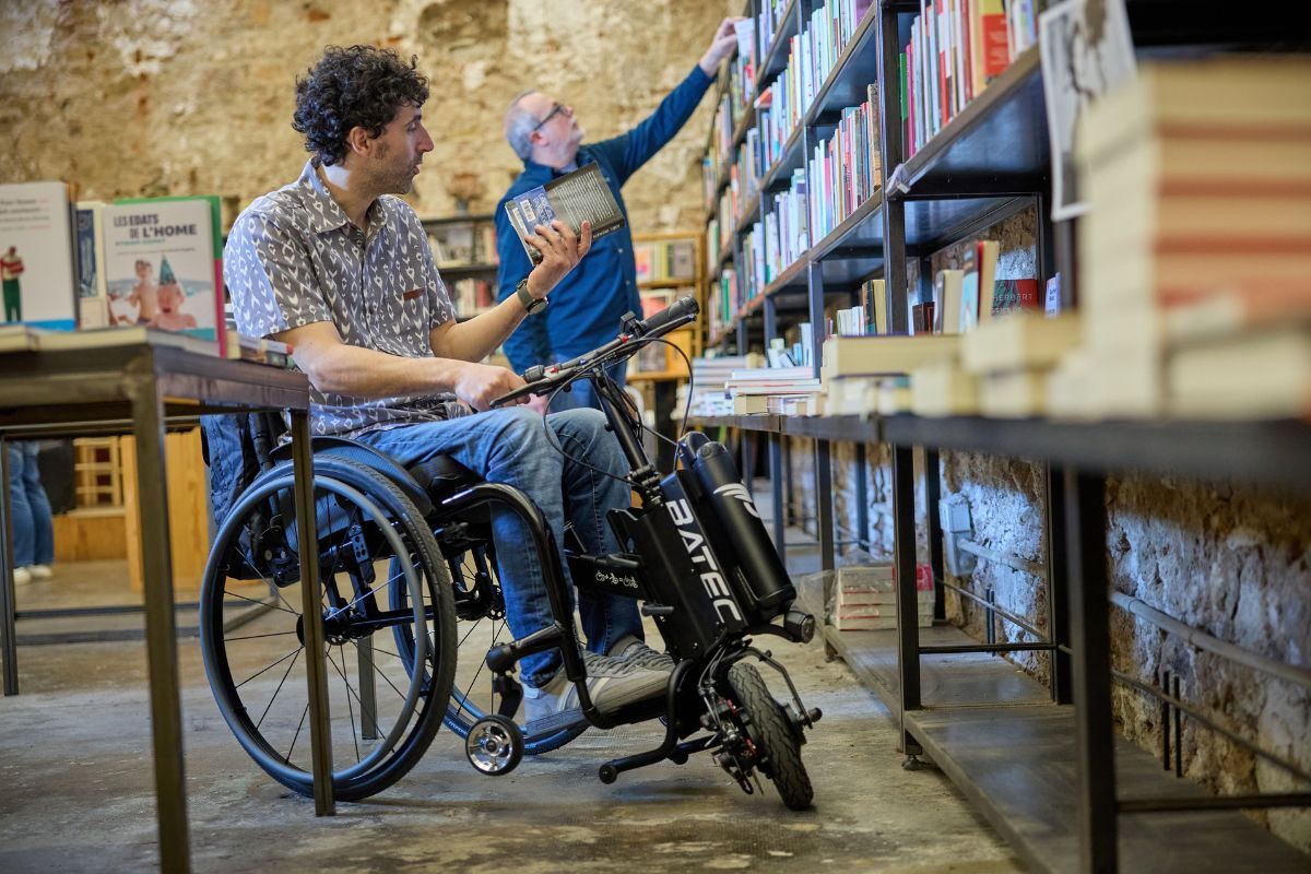 Man in wheelchair at bookstore, looking at books. Another man reaches for a book on a shelf.