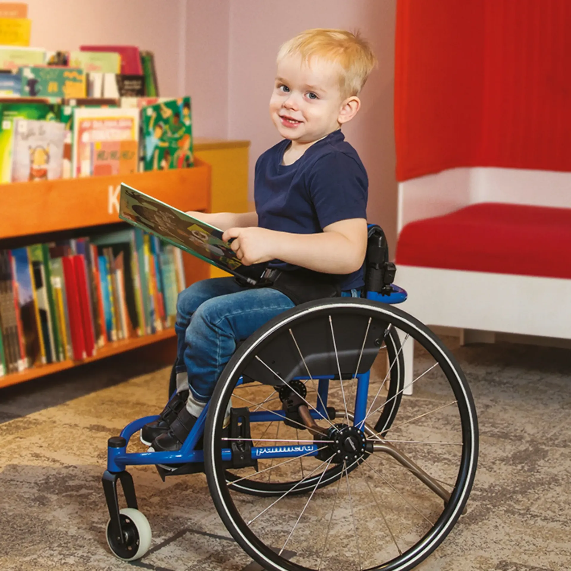 Young child in a blue wheelchair reads a book in a library, smiling, shelves of books in background.