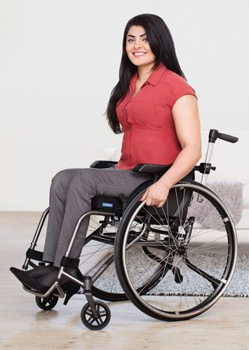 Woman in wheelchair, wearing a red top and gray pants, smiling, indoors.