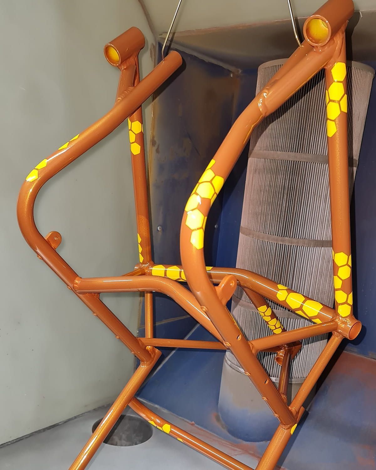 Brown metal wheelchair frame with yellow honeycomb details, hanging in a spray booth.