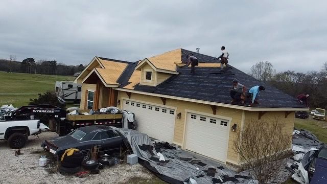 Storm-damaged house with workers on roof, overturned cars, and debris scattered across the driveway