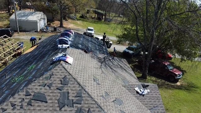 Workers repairing a gray shingled roof beside trees and a red car in a suburban yard