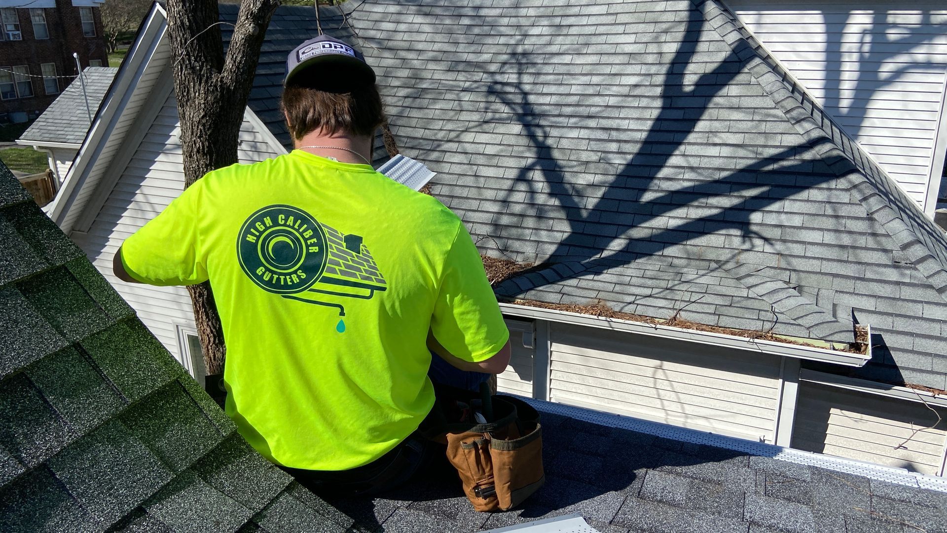 Worker in neon yellow shirt and black cap repairs a shingled roof near a dormer window.