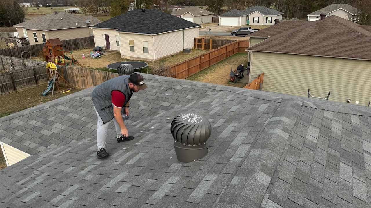 Worker on a gray shingle roof using a hand tool near a spinning roof turbine vent.