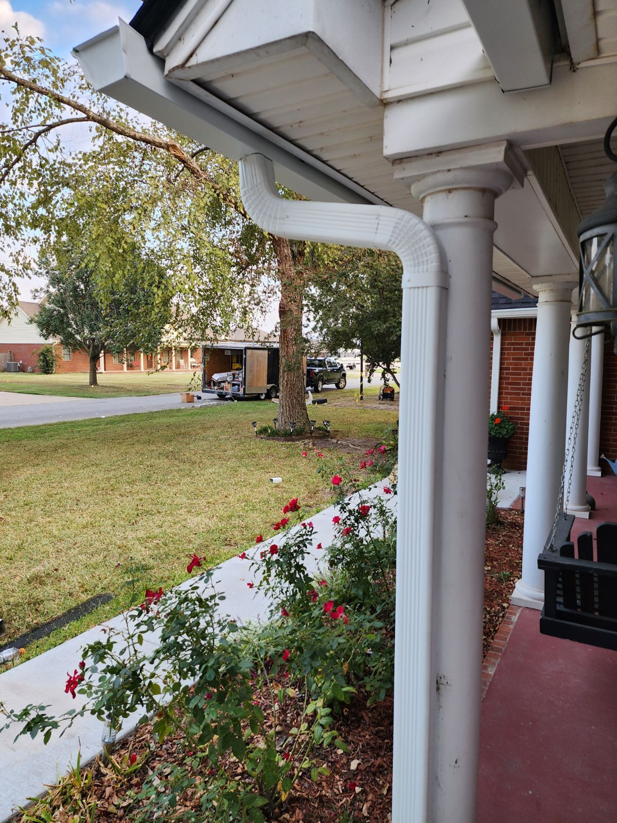 Front porch with white columns, red floor, flower bed, and a grassy yard with trees and a street beyond.