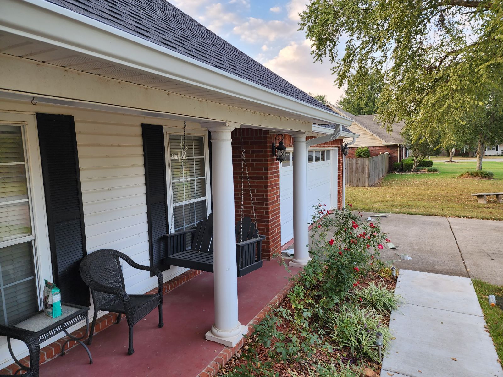 Front porch with white columns, black chairs, flower bed, and sidewalk beside a brick-and-siding house