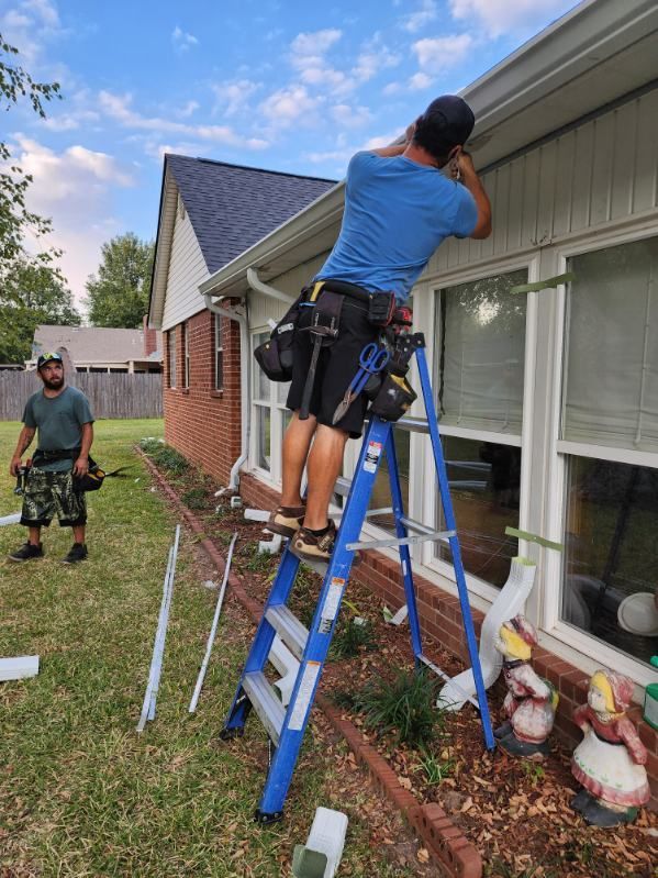 Person on a blue ladder cleaning gutters beside a house while another stands nearby in the yard.